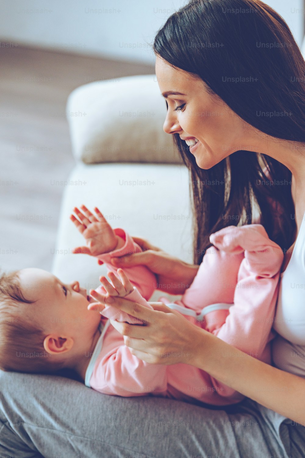 Side view of cheerful beautiful young woman holding baby girl on her knees and looking at her with love while sitting on the couch at home