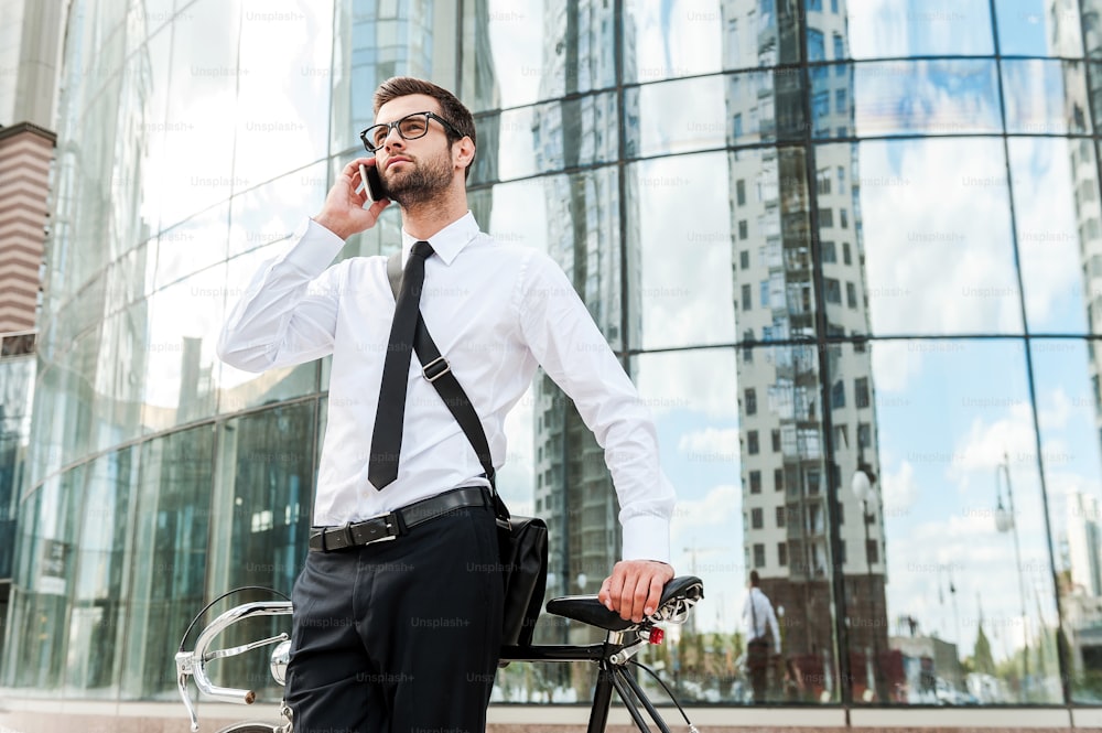 Low angle view of handsome young businessman talking on the mobile phone while leaning at the bicycle