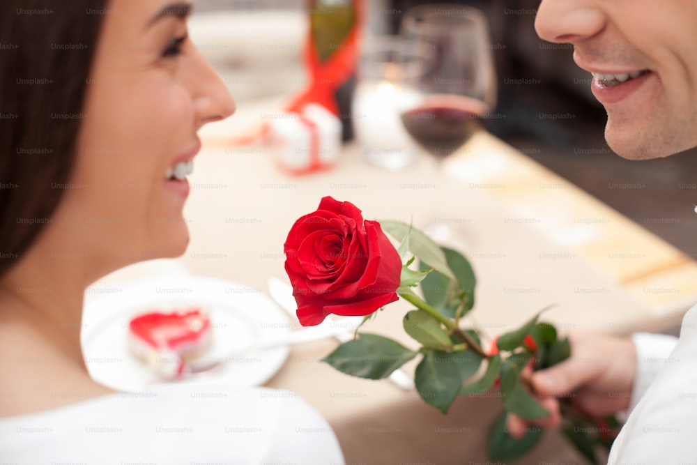 Close up of beautiful young loving couple celebrating Valentine day in cafe. The man is giving to a woman a rose. They are sitting at the table and smiling