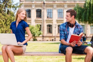 Handsome young man sitting on the bench and reading book while beautiful woman sitting near him and using computer