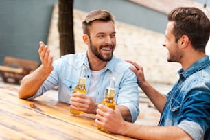 Two cheerful young men talking to each other and holding bottles with beer while standing outdoors