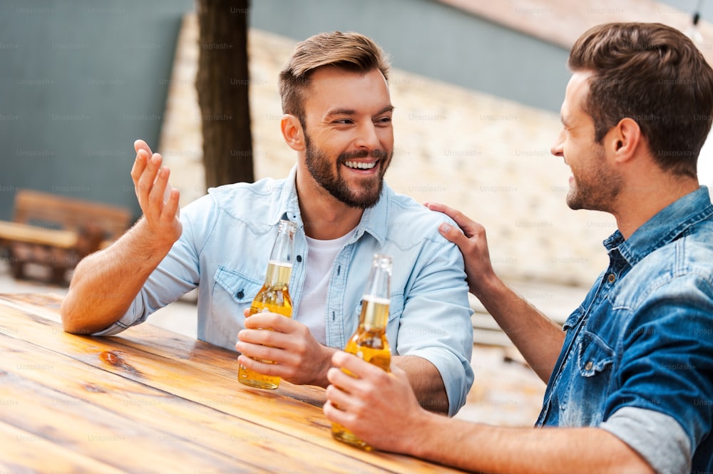 Two cheerful young men talking to each other and holding bottles with beer while standing outdoors