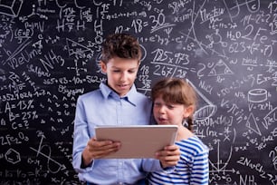 Niño y niña lindos con tableta en la escuela frente a una pizarra grande. Foto de estudio sobre fondo negro.
