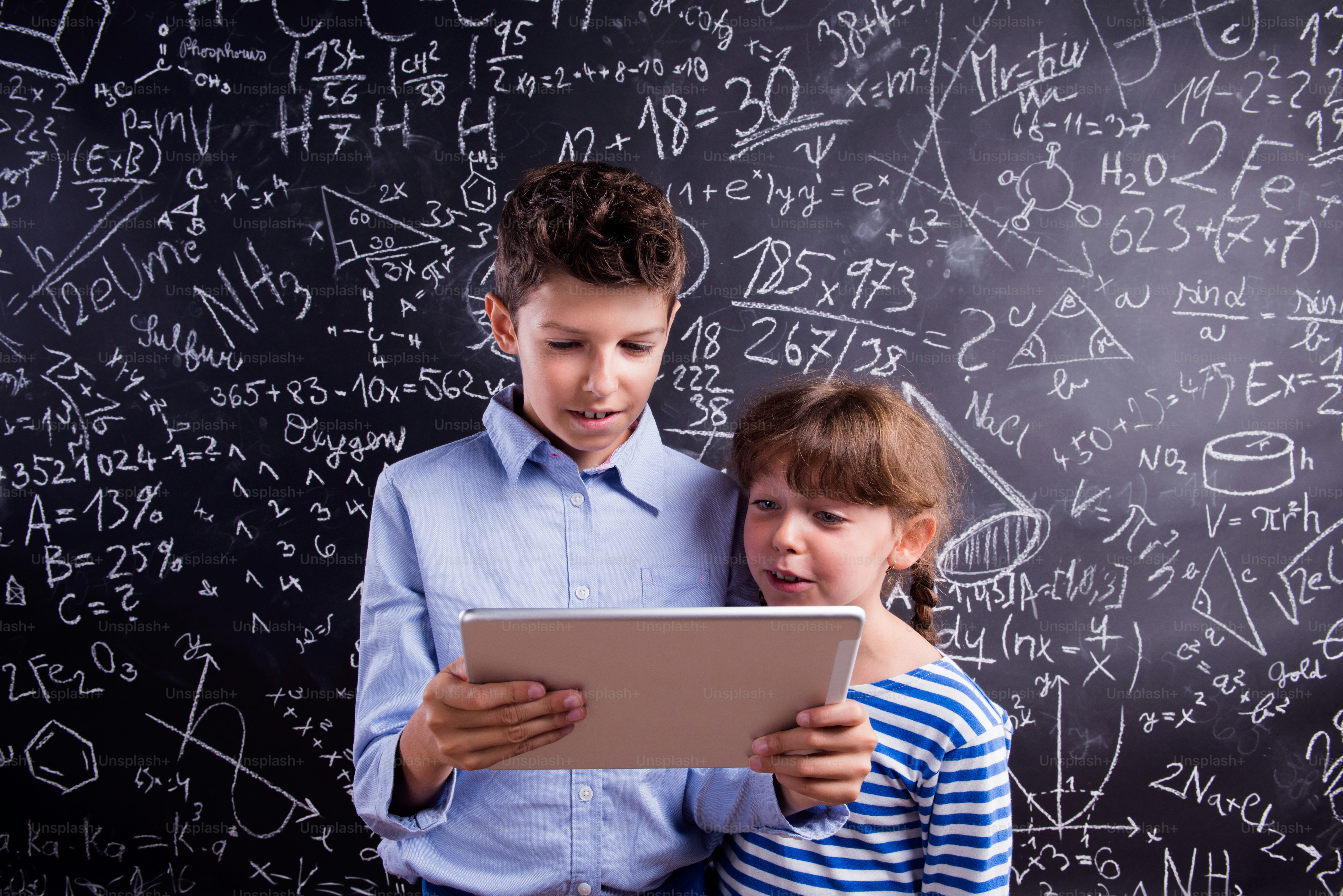 Cute boy and girl with tablet at school in front of a big blackboard. Studio shot on black background