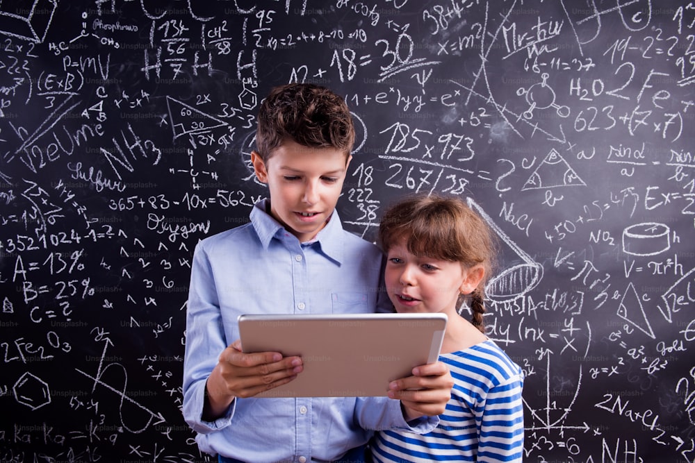 Cute boy and girl with tablet at school in front of a big blackboard. Studio shot on black background.