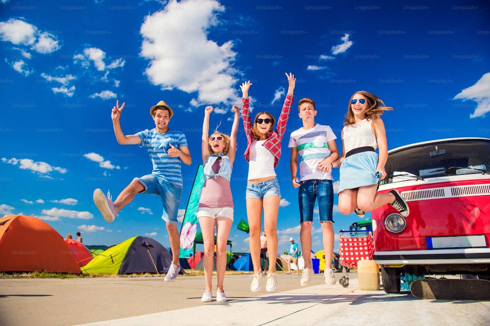 Group of teenage boys and girls at summer music festival jumping by vintage red campervan