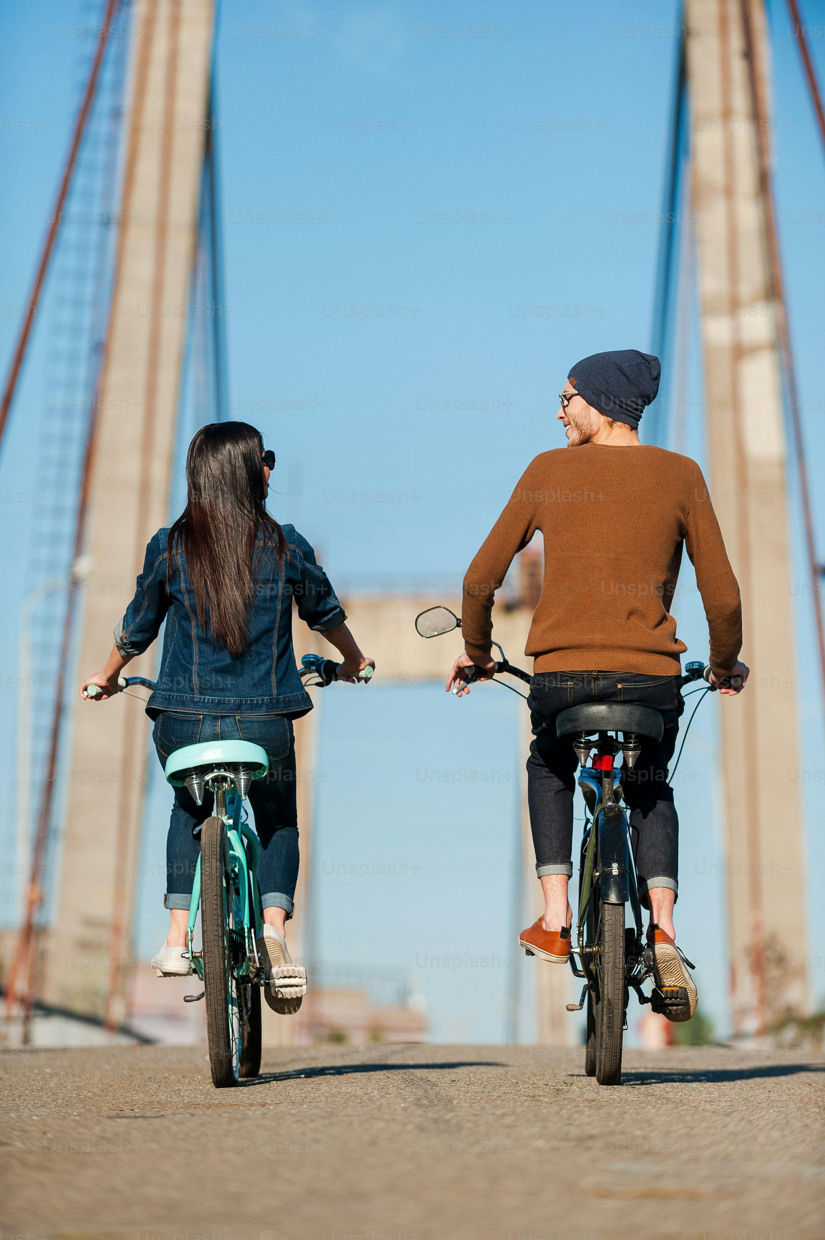 Rear view of beautiful young couple riding bicycles along the bridge and looking at each other