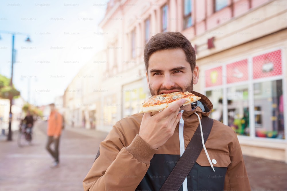 Handsome young man eating a slice of pizza outside on the street