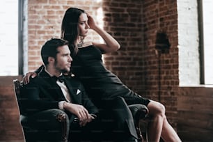 Schönes junges Paar in formeller Kleidung schaut zum Fenster, während es zusammen auf einem Stuhl sitzt