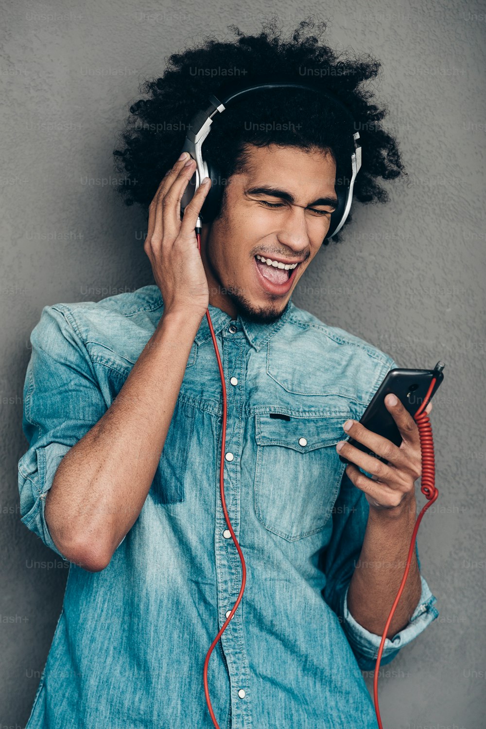 Joven africano usando audífonos y sosteniendo su teléfono inteligente mientras mantiene los ojos cerrados y la boca abierta contra el fondo gris