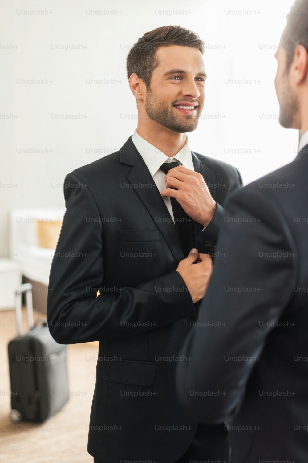 Jovem bonito em traje formal ajustando sua gravata e sorrindo enquanto está de pé contra o espelho no quarto de hotel