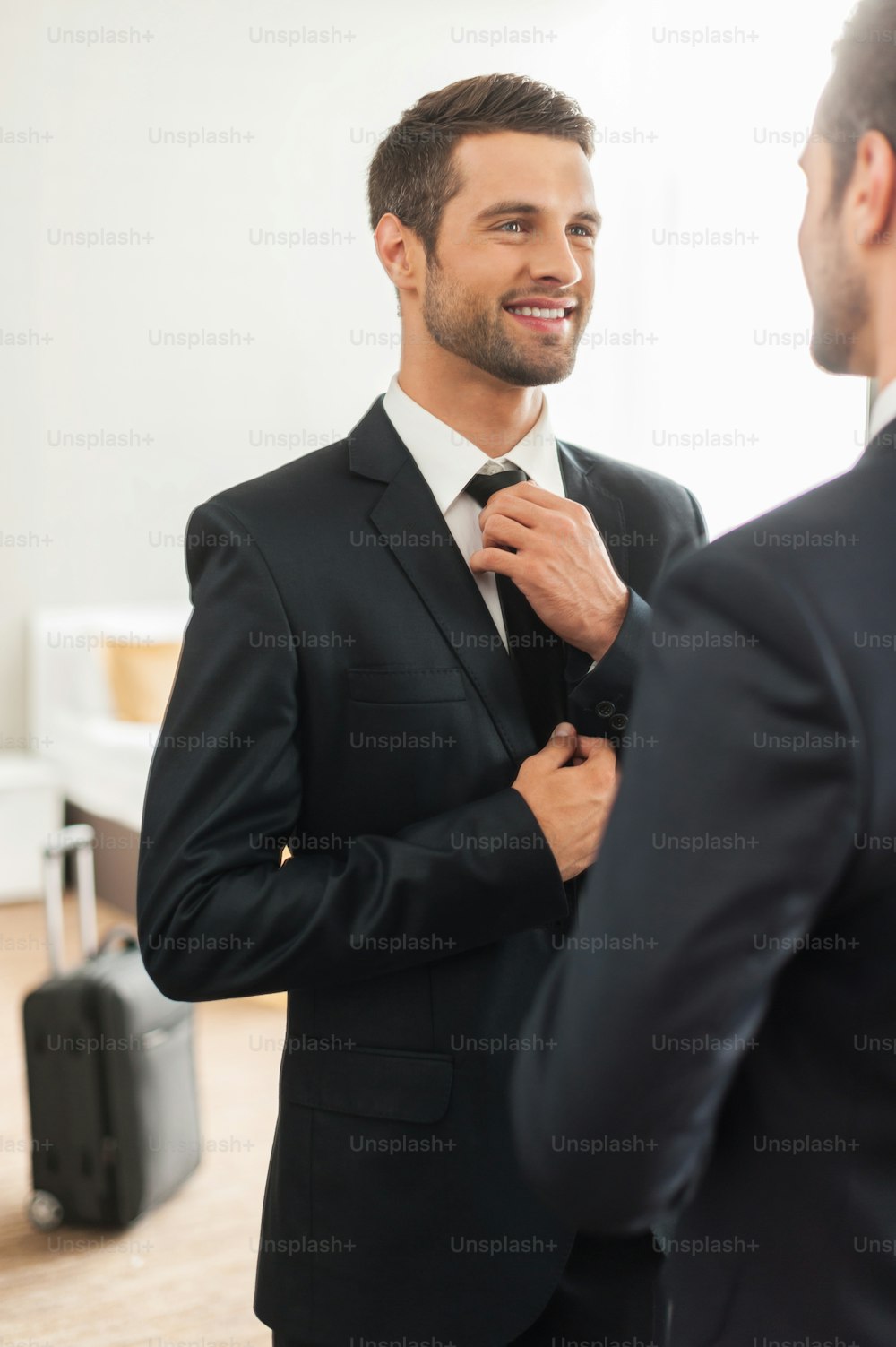 Handsome young man in formalwear adjusting his necktie and smiling while standing against mirror in hotel room