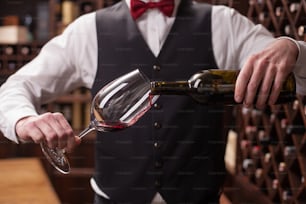 Primo piano delle mani di un giovane sommelier che versa vino rosso dalla bottiglia al bicchiere. È in piedi in una cantina