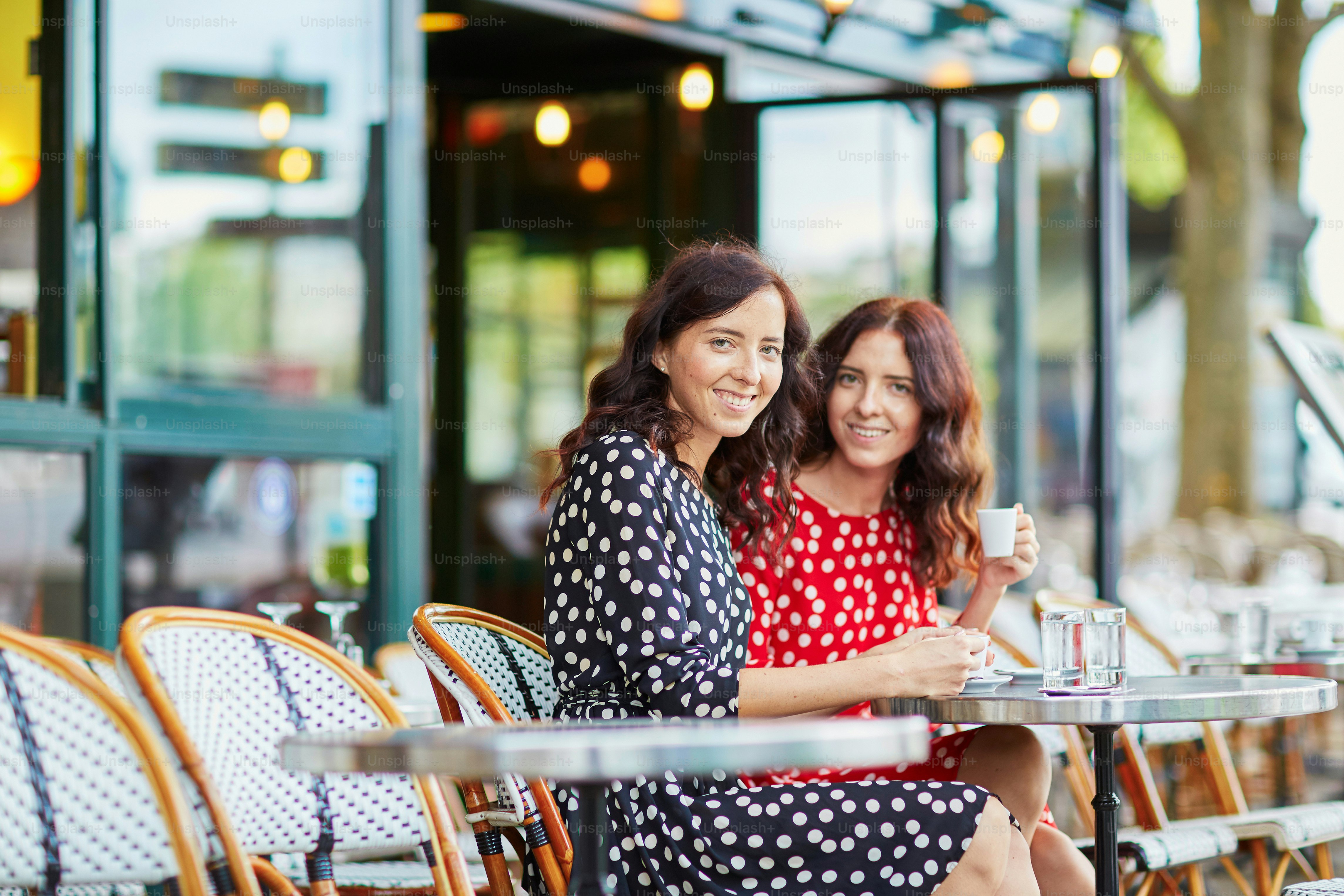 Beautiful twin sisters drinking coffee in a cozy outdoor cafe in Paris, France. Happy smiling girls enjoy their vacation in Europe
