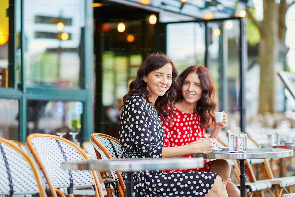 フランス・パリの居心地の良い屋外カフェでコーヒーを飲む美しい双子の姉妹。幸せな笑顔の女の子は、ヨーロッパでの休暇を楽しんでいます