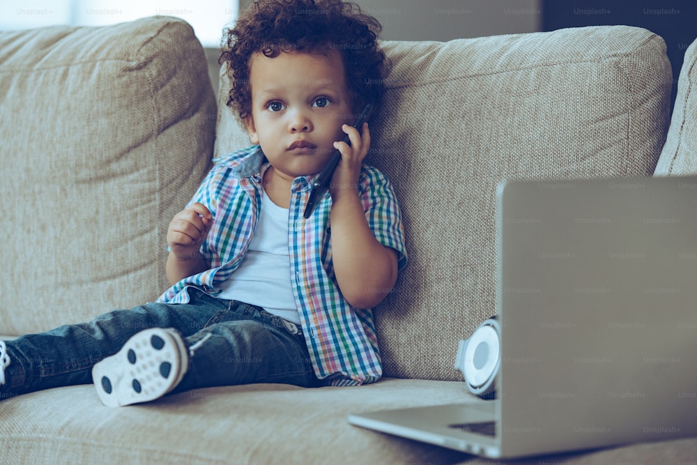 Piccolo bambino africano che parla al telefono cellulare e guarda la macchina fotografica mentre è seduto sul divano di casa