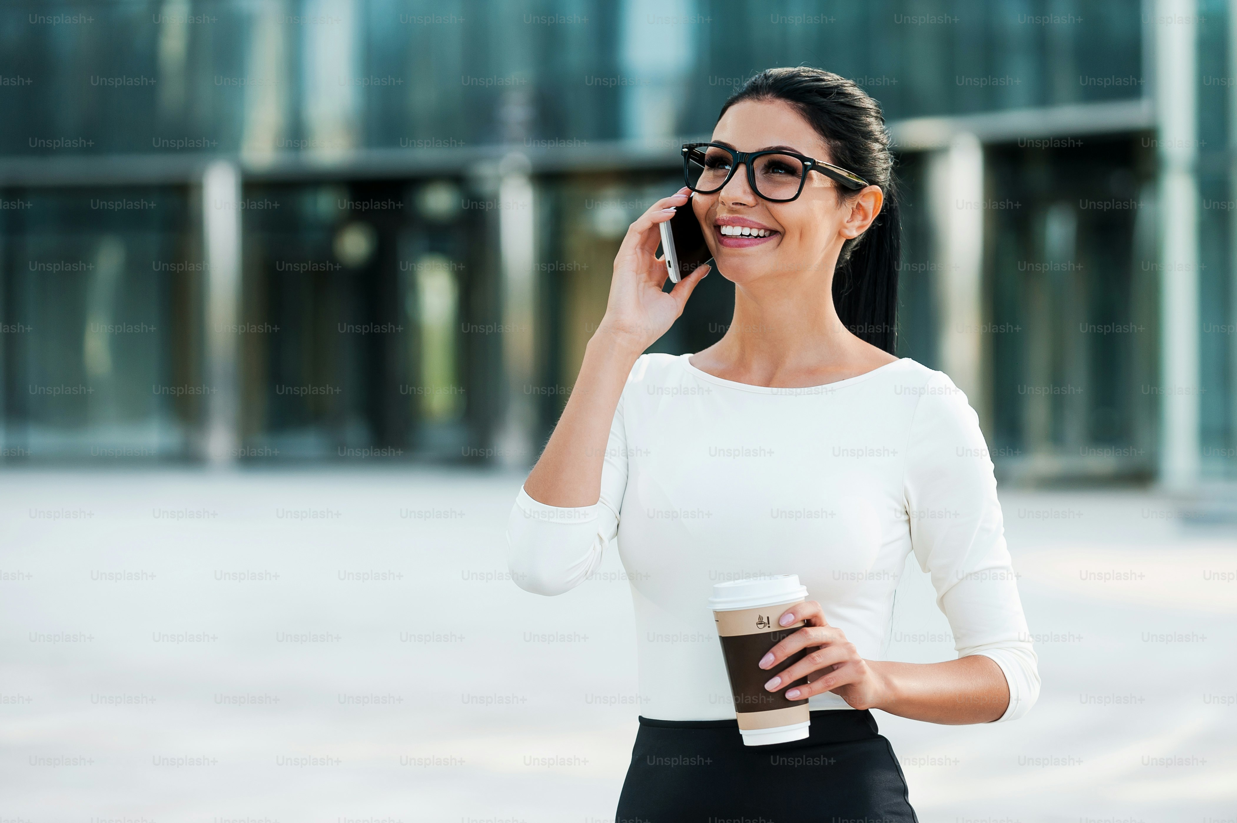 Cheerful young businesswoman talking on the mobile phone and holding cup of coffee while standing outdoors