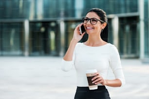 Cheerful young businesswoman talking on the mobile phone and holding cup of coffee while standing outdoors