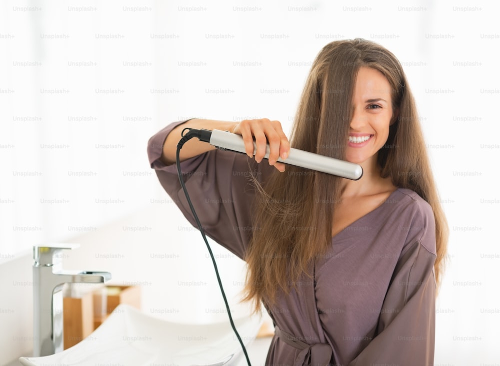 Glückliche junge Frau glättet Haare im Badezimmer