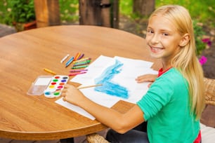 Vue de dessus d’une jolie petite fille dessinant quelque chose sur papier et souriant tout en étant assise à la table et à l’extérieur