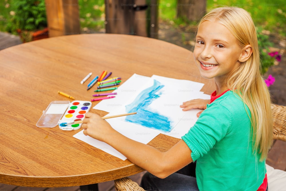 Top view of cute little girl drawing something on paper and smiling while sitting at the table and outdoors