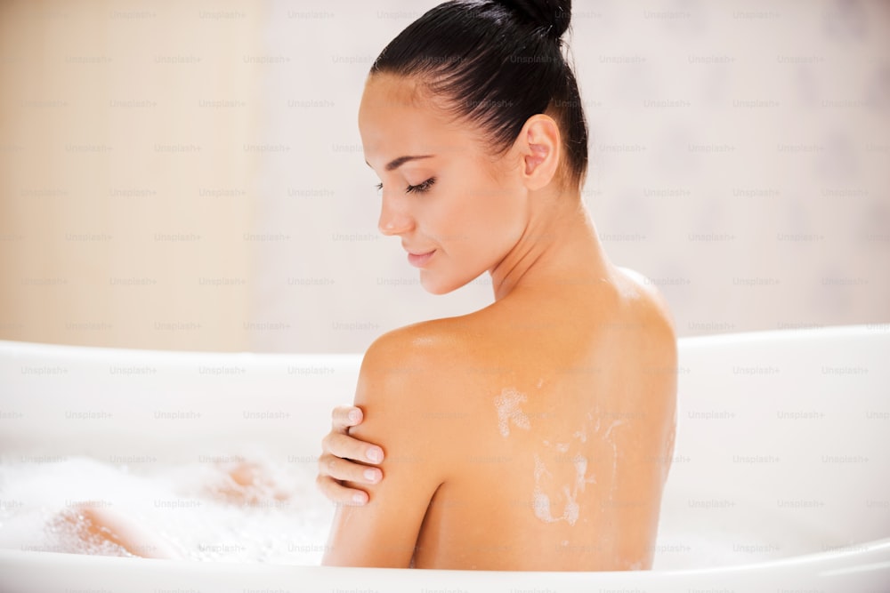 Vue arrière d’une jeune femme séduisante touchant son épaule tout en profitant d’un bain moussant