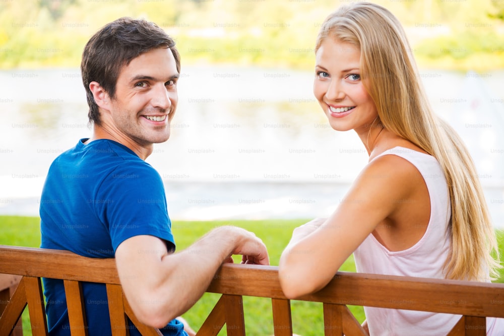 Rear view of beautiful young loving couple sitting on the bench together and looking over shoulder with smile