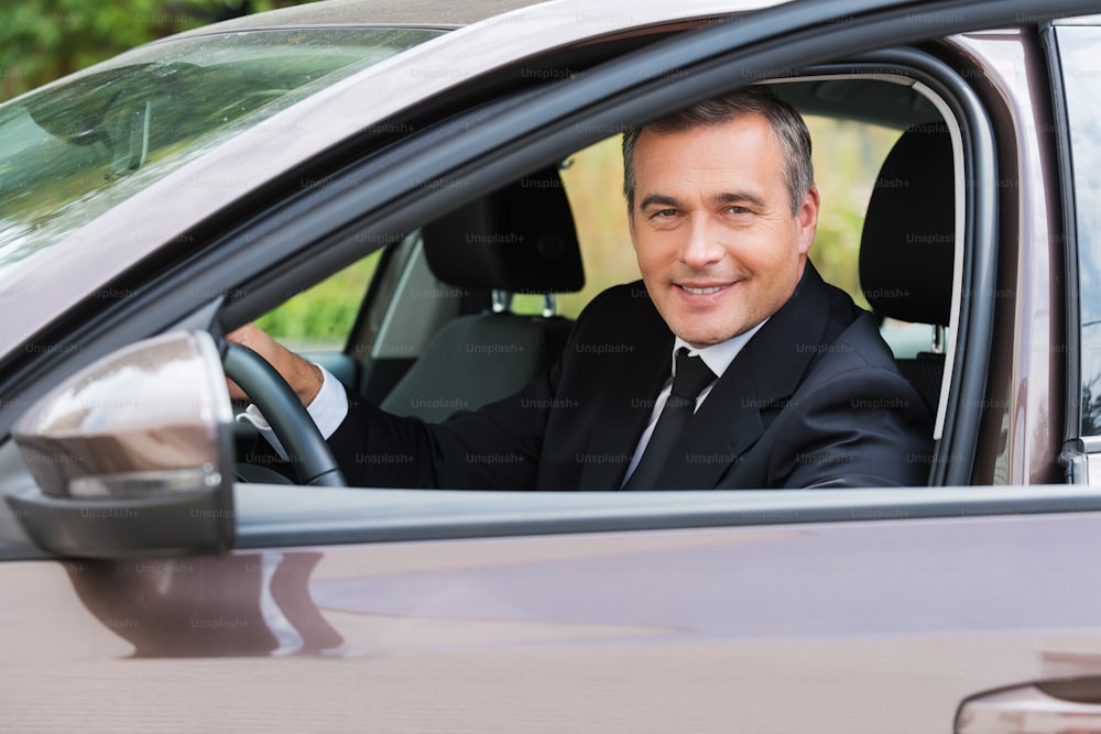 Cheerful mature man in formalwear looking at car and smiling while sitting in car