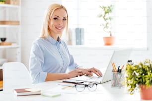 Cheerful young beautiful woman working on laptop and looking at camera with smile while sitting at her working place