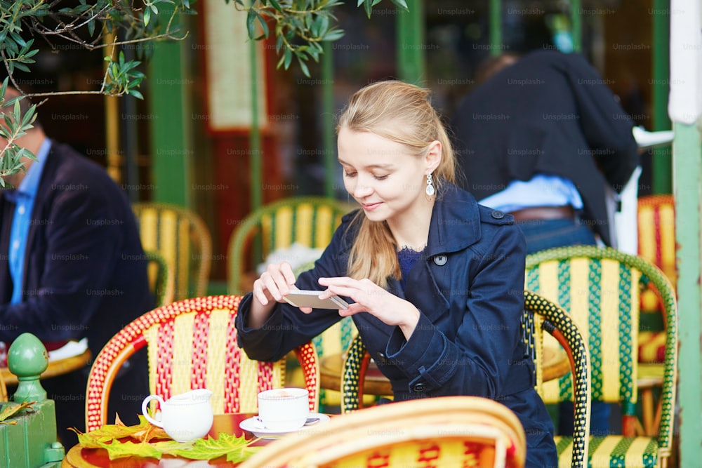 Bella giovane donna che beve cioccolata calda nel caffè all'aperto parigino e scatta foto con il suo telefono cellulare