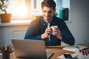 Jeune homme joyeux et beau tenant une tasse de café et regardant son ordinateur portable avec le sourire tout en étant assis à son lieu de travail