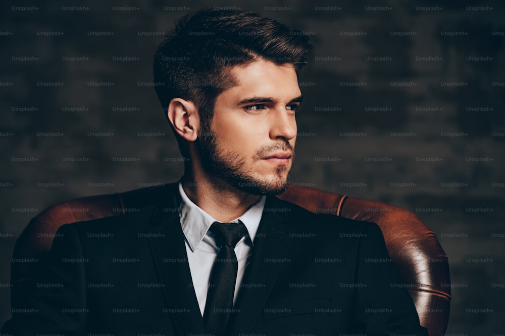 Portrait of young handsome man in suit holding his sunglasses and looking at camera while sitting in leather chair against dark grey background