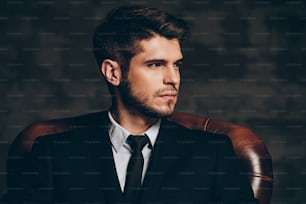Ritratto di giovane bell'uomo in giacca e cravatta che tiene i suoi occhiali da sole e guarda la macchina fotografica mentre è seduto in una sedia di pelle su sfondo grigio scuro