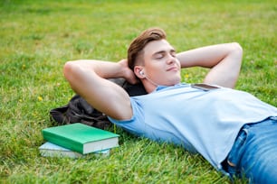 Handsome young man holding hands behind head and smiling while lying on grass and listening to MP3 Player
