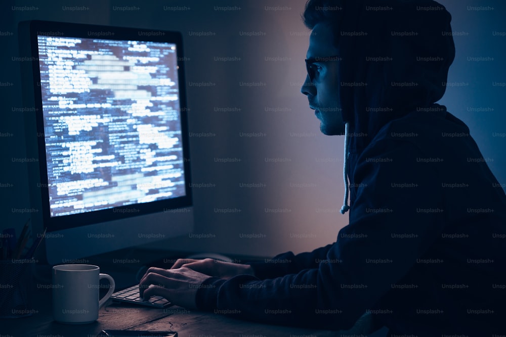 Vista lateral de un joven que escribe y mira el monitor de la computadora mientras está sentado en la mesa en el cuarto oscuro