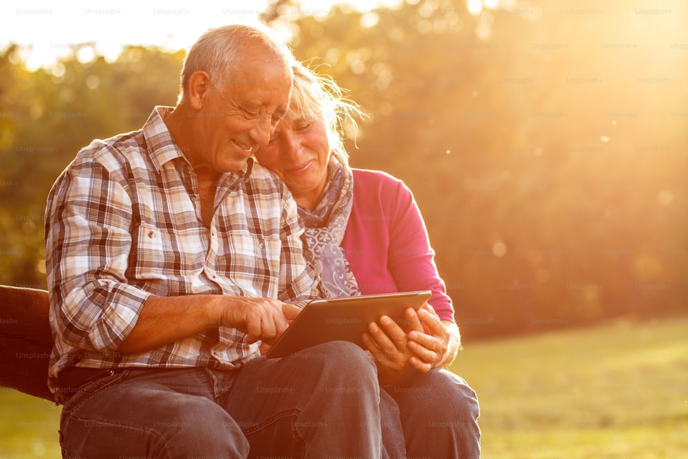 Senior couple sitting on a park bench on sunny autumn day with tablet and relaxing.