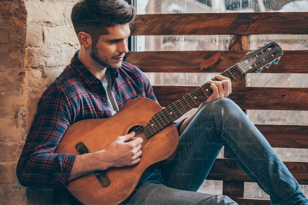 Hübscher junger Mann, der Gitarre spielt, während er auf der Fensterbank sitzt