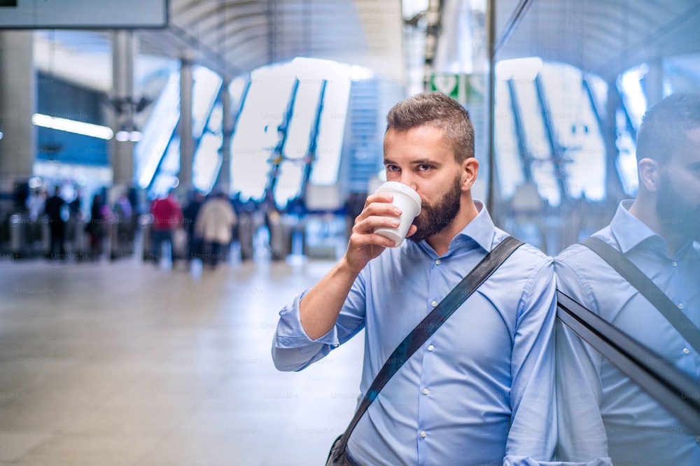 Close up of handsome hipster businessman in blue shirt drinking coffee, standing on subway station
