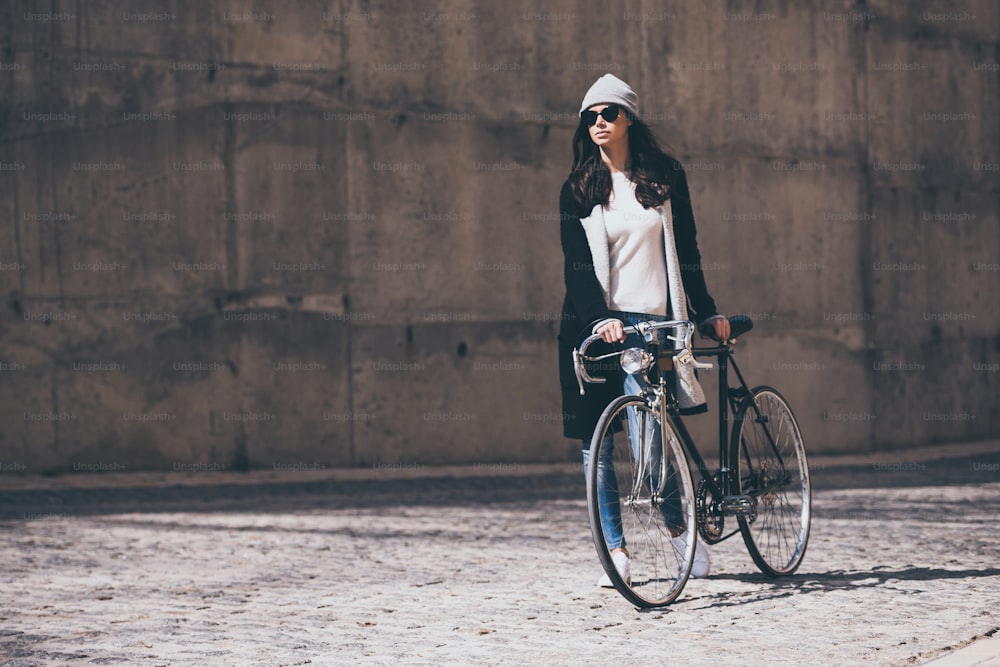 Beautiful young woman in sunglasses rolling her bicycle and looking away while walking outdoors