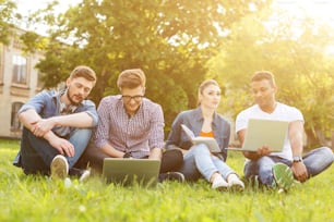 Glückliche junge Studenten entspannen sich auf Gras in der Nähe einer Universität. Sie sitzen und reden. Männer benutzen Laptops und lächeln. Frau liest ein Buch