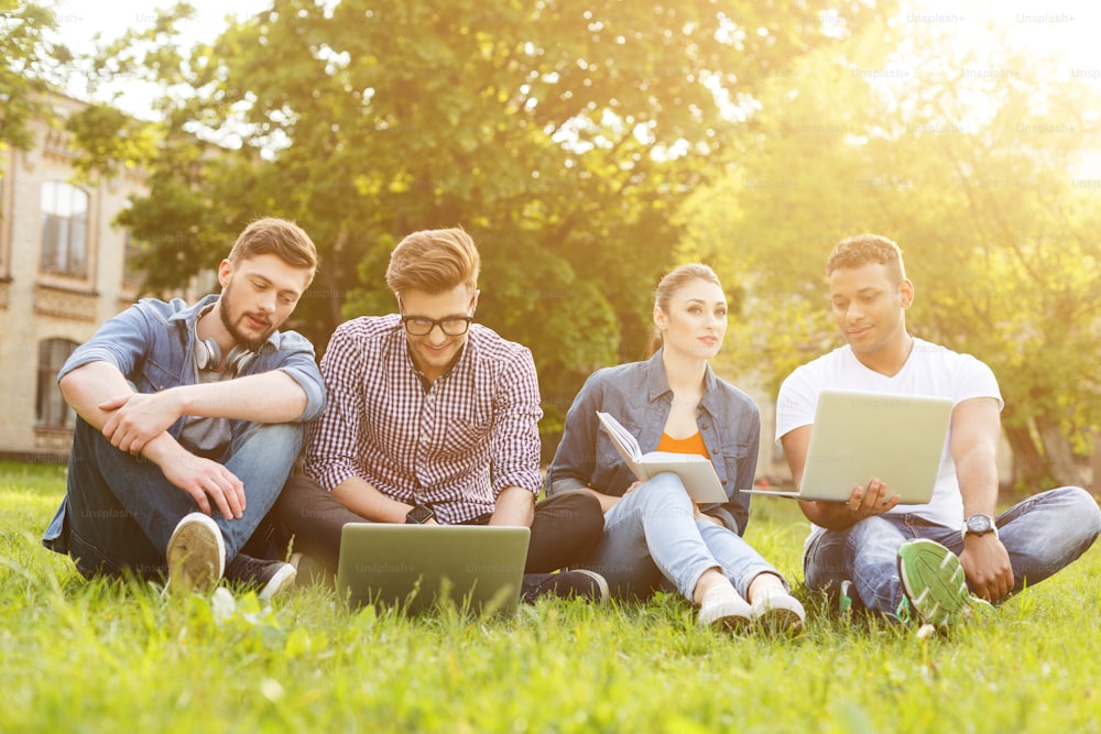 Happy young students are relaxing on grass near a university. They are sitting and talking. Men are using laptops and smiling. Woman is reading a book