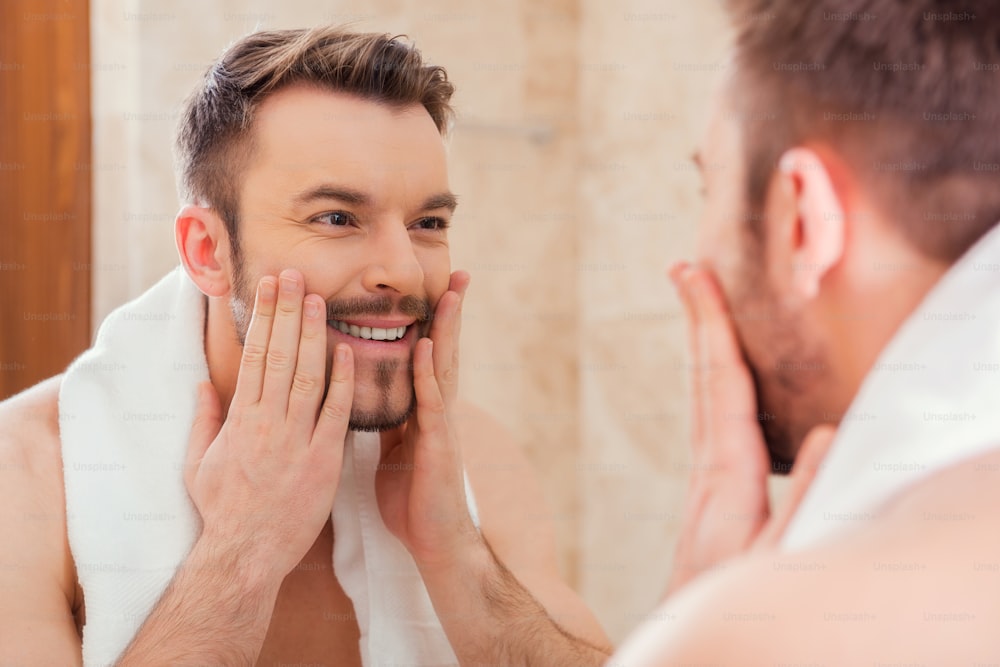 Handsome young man touching his face and smiling while standing in front of the mirror