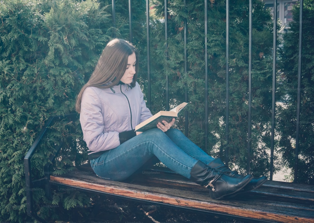 Giovane ragazza adolescente seduta su una panchina del parco che legge un libro affascinante