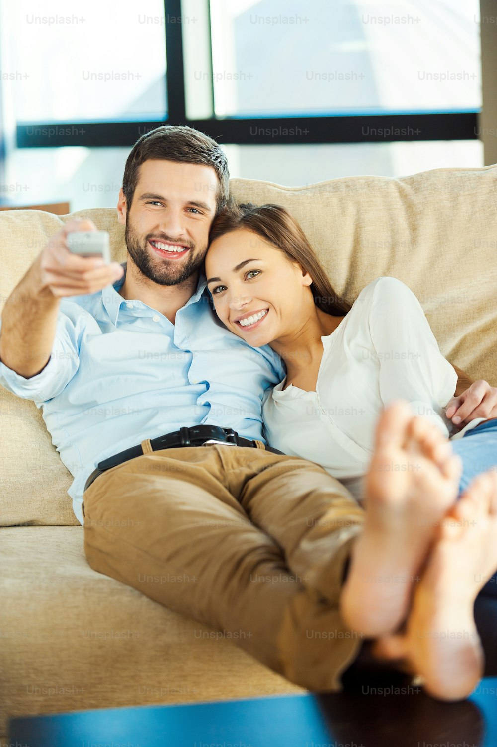 ソファに一緒に座ってテレビを見ている美しい若い愛情のあるカップルがリモコンを持って微笑んでいる間