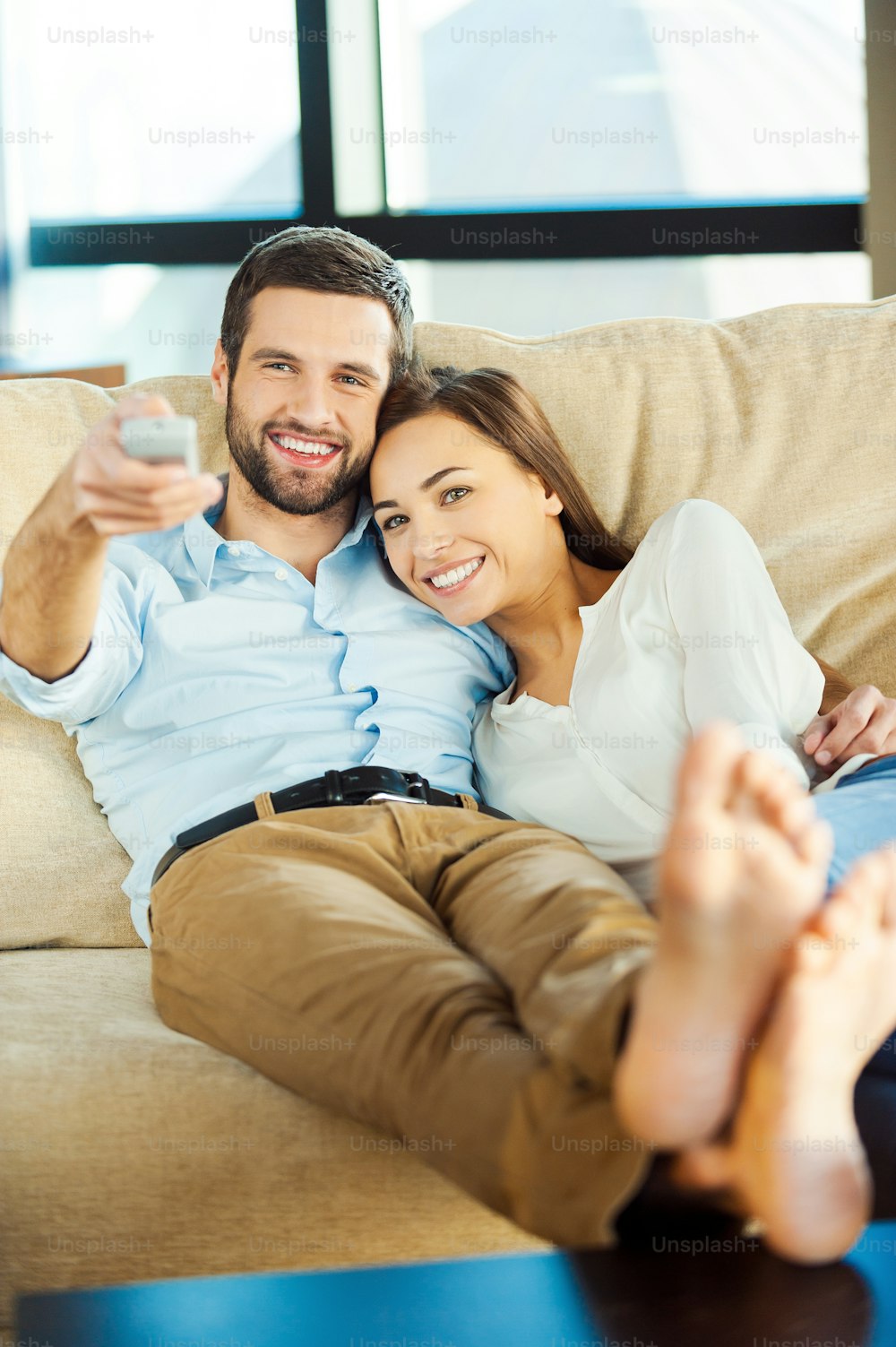 Schönes junges Liebespaar, das zusammen auf der Couch sitzt und fernsieht, während der Mann die Fernbedienung hält und lächelt