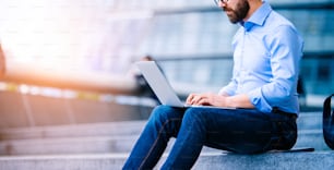 Unrecognizable hipster manager sitting on stairs on sunny day, working on laptop, London, City Hall