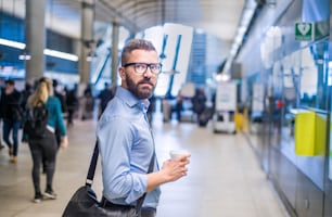 Empresário hipster bonito em camisa azul segurando uma xícara de café, de pé na estação de metrô