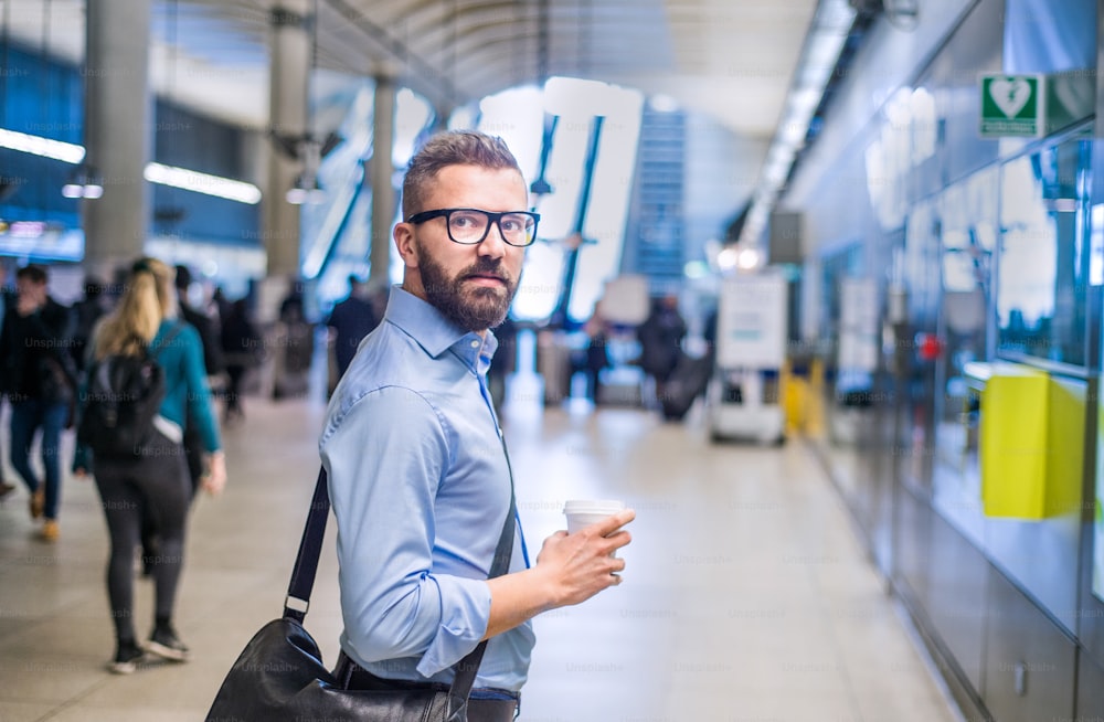 Hübscher Hipster-Geschäftsmann im blauen Hemd mit Kaffeetasse in der Hand, auf U-Bahn-Station stehend