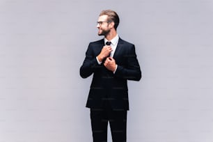 Studio shot of handsome young man in full suit adjusting his necktie and smiling