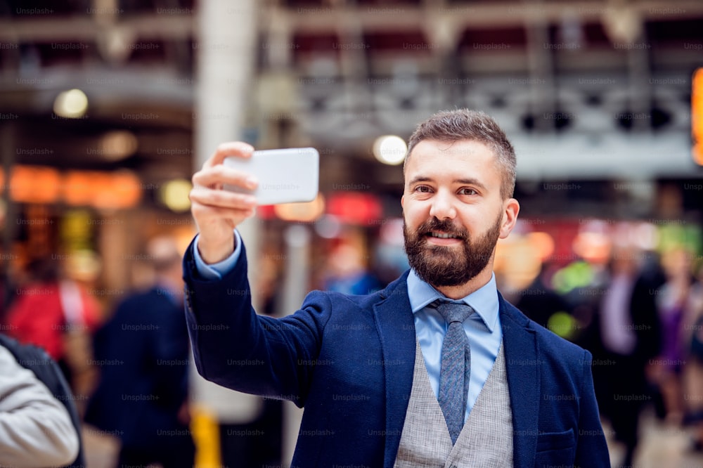 Hombre de negocios hipster en traje con teléfono inteligente tomando selfie, estación de tren de Londres abarrotada